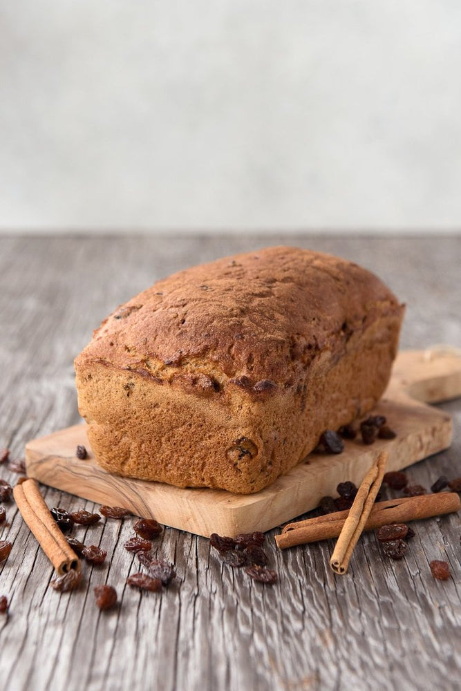 whole loaf gluten free cinnamon raisin bread on cutting board displayed with cinnamon sticks & raisins